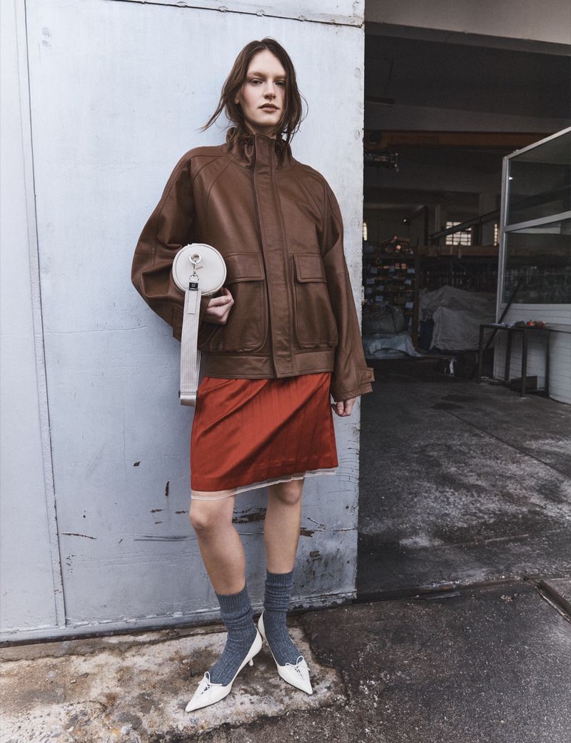 Model in oversized brown leather jacket and reddish-orange skirt holding a small white handbag against a pale industrial backdrop.
