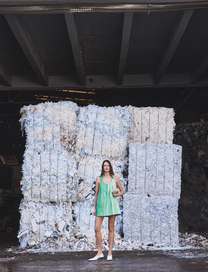 Model wearing a green mini dress standing before stacked bales of shredded white paper in an industrial warehouse.