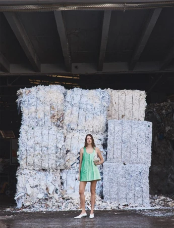 Model wearing a green mini dress standing before stacked bales of shredded white paper in an industrial warehouse.