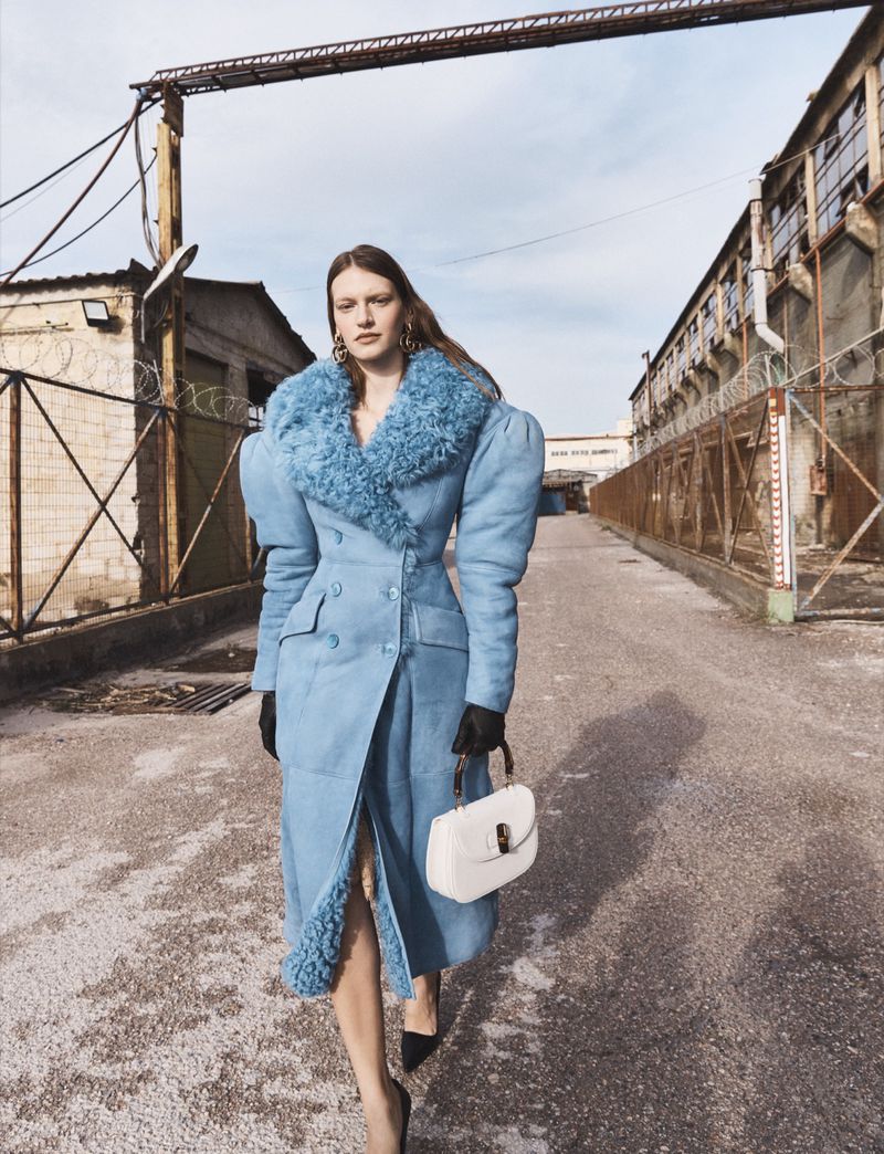 Model wearing a blue coat with fur collar and puffed shoulders holding a white handbag on an industrial road.