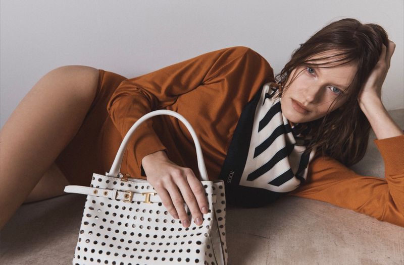 Woman in a brown shirt lying down, holding a white, perforated handbag with a striped scarf, indoor in a studio setting.
