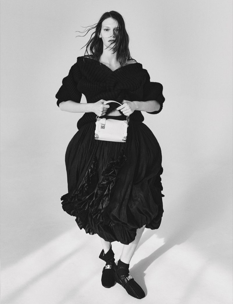 Model in voluminous black outfit holding a small white handbag against a plain background