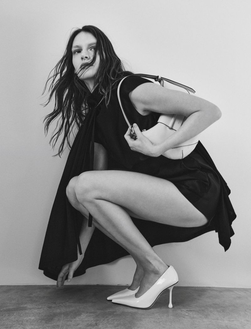 Model in black dress holding a white handbag and wearing white heels against a plain studio background.