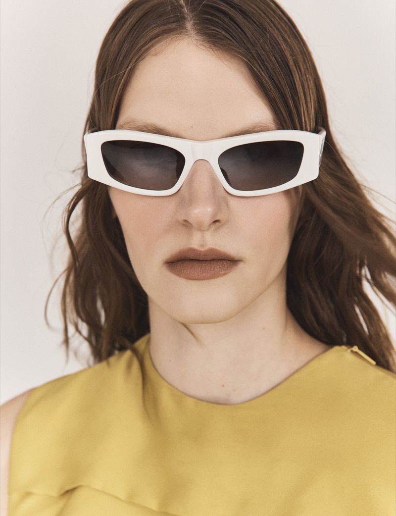 Model wearing thick white framed sunglasses and a mustard top against a plain studio background for Vogue Greece.
