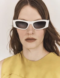 Model wearing thick white framed sunglasses and a mustard top against a plain studio background for Vogue Greece.