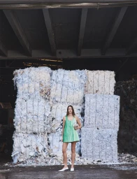 Model wearing a green mini dress standing before stacked bales of shredded white paper in an industrial warehouse.