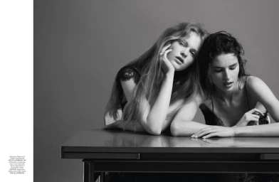 Two female models posing seated at a table against a plain grey studio backdrop in a black and white portrait photograph.