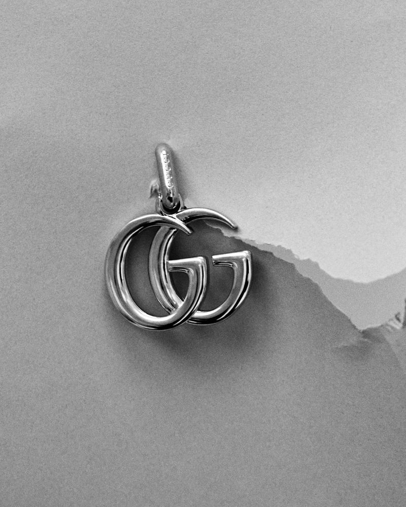 Silver double-G logo pendant against a textured grey paper background