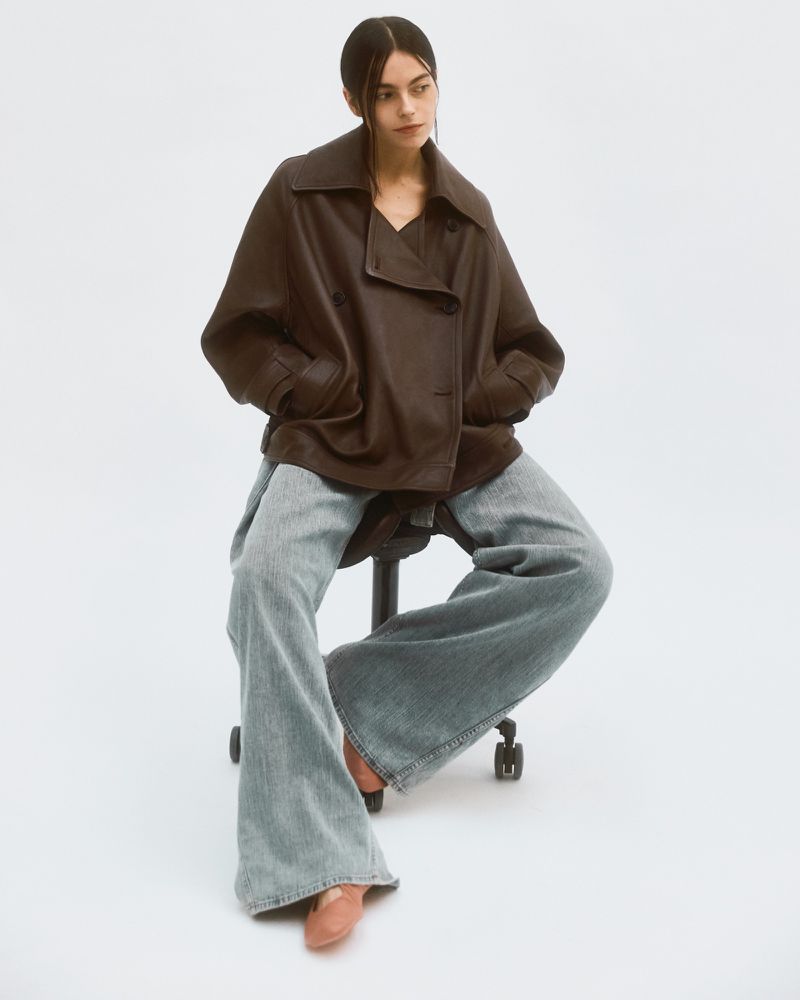 Model seated on a wheeled stool wearing an oversized brown leather jacket and wide-leg denim jeans against a white background