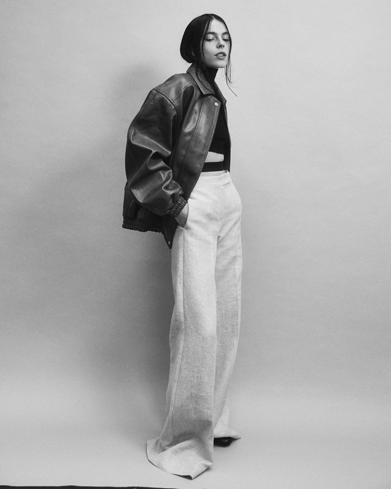 Model in oversized leather jacket and light trousers standing against grey studio backdrop
