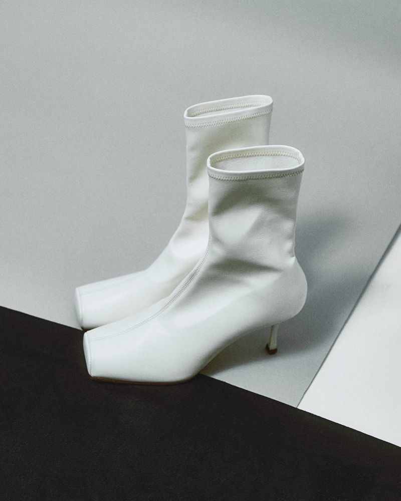 White ankle boots with square toes and kitten heels posed against a geometric backdrop of grey and black planes.
