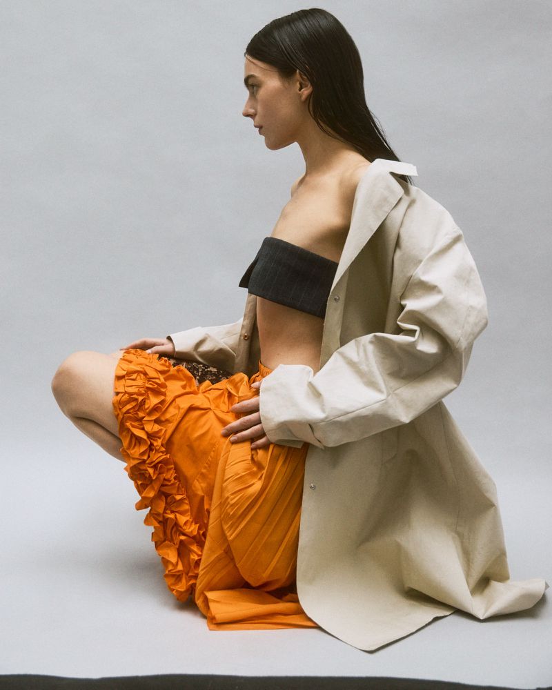 Profile view of a model crouching, wearing an orange ruffled skirt, dark bandeau top, and an oversized beige coat against a white background.