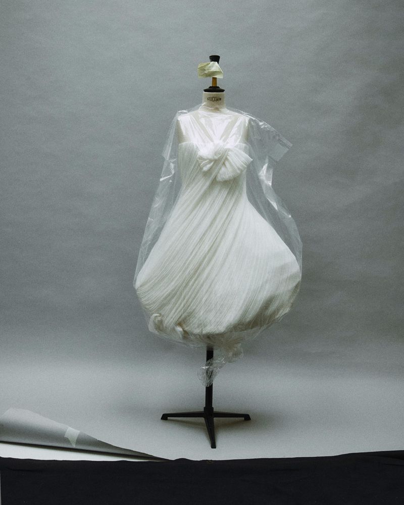 White dress on a dressmaker mannequin covered in clear plastic film against a grey studio backdrop.