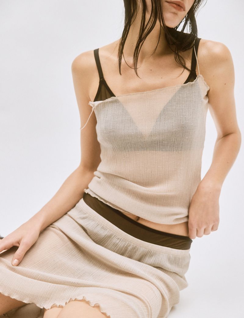 Model wearing a sheer cream top and matching skirt over dark undergarments against a plain white studio background.