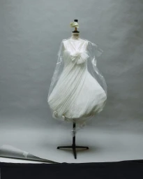 White dress on a dressmaker mannequin covered in clear plastic film against a grey studio backdrop.