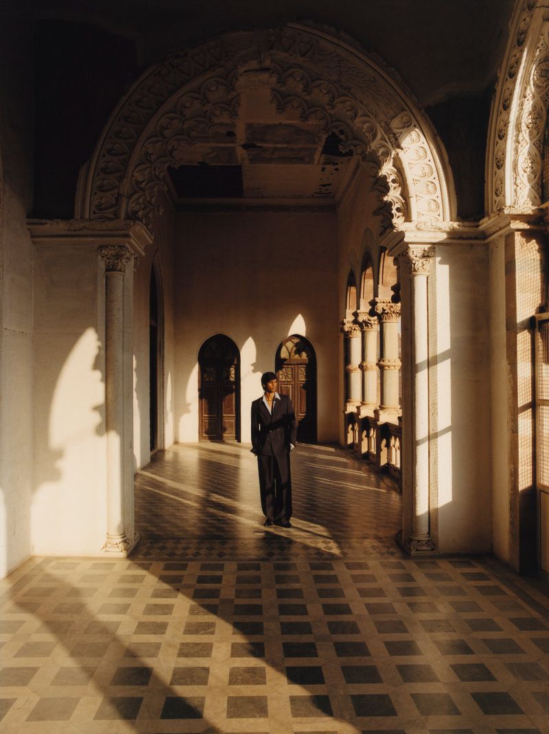 Model standing in a grand historical hall with arched colonnades and patterned tile flooring under warm, directional sunlight.