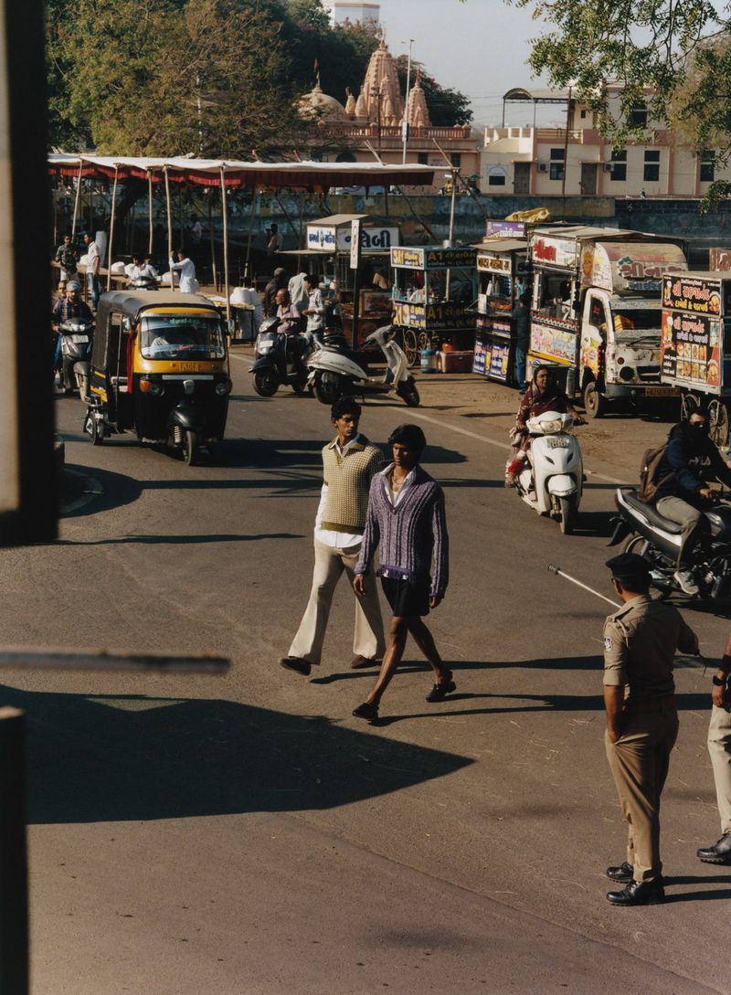 Two men walking across a busy sunny street with autorickshaws, scooters, and food stalls in the background