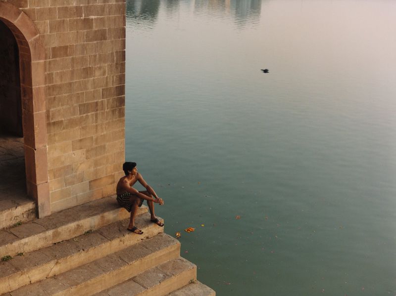 Man seated on stone steps beside a calm lake with an arched brick structure in the foreground