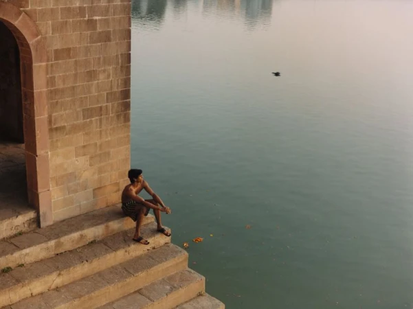 Man seated on stone steps beside a calm lake with an arched brick structure in the foreground