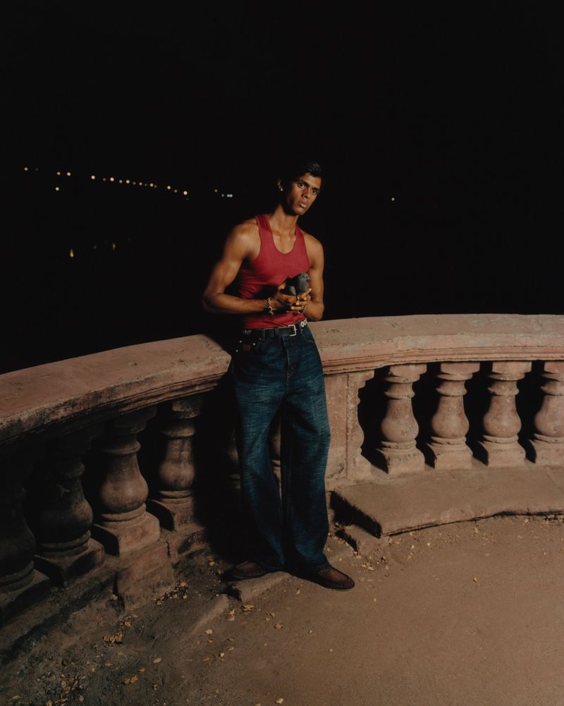 A man wearing a red tank top and blue jeans leaning against a stone balustrade at night.