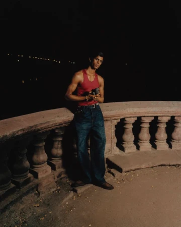 A man wearing a red tank top and blue jeans leaning against a stone balustrade at night.