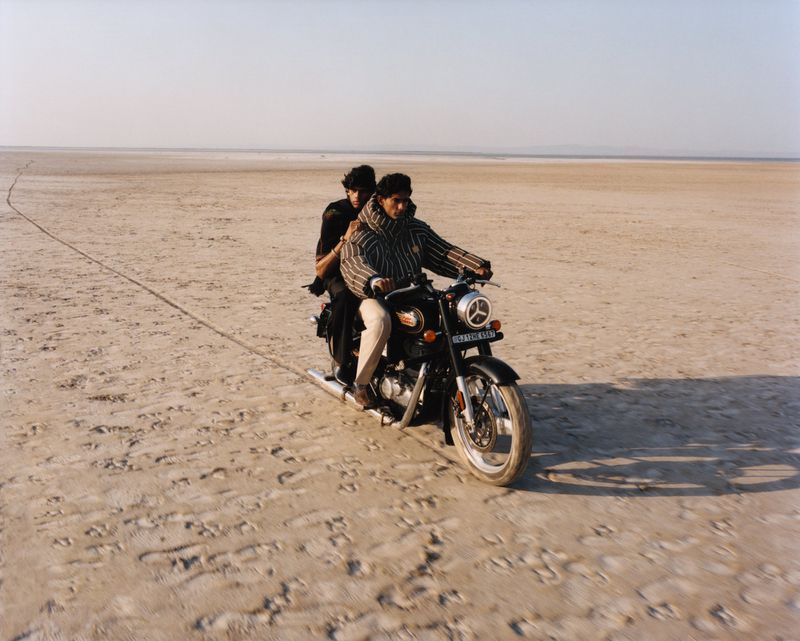 Two men riding a motorcycle across a vast empty desert salt flat under a clear sky.