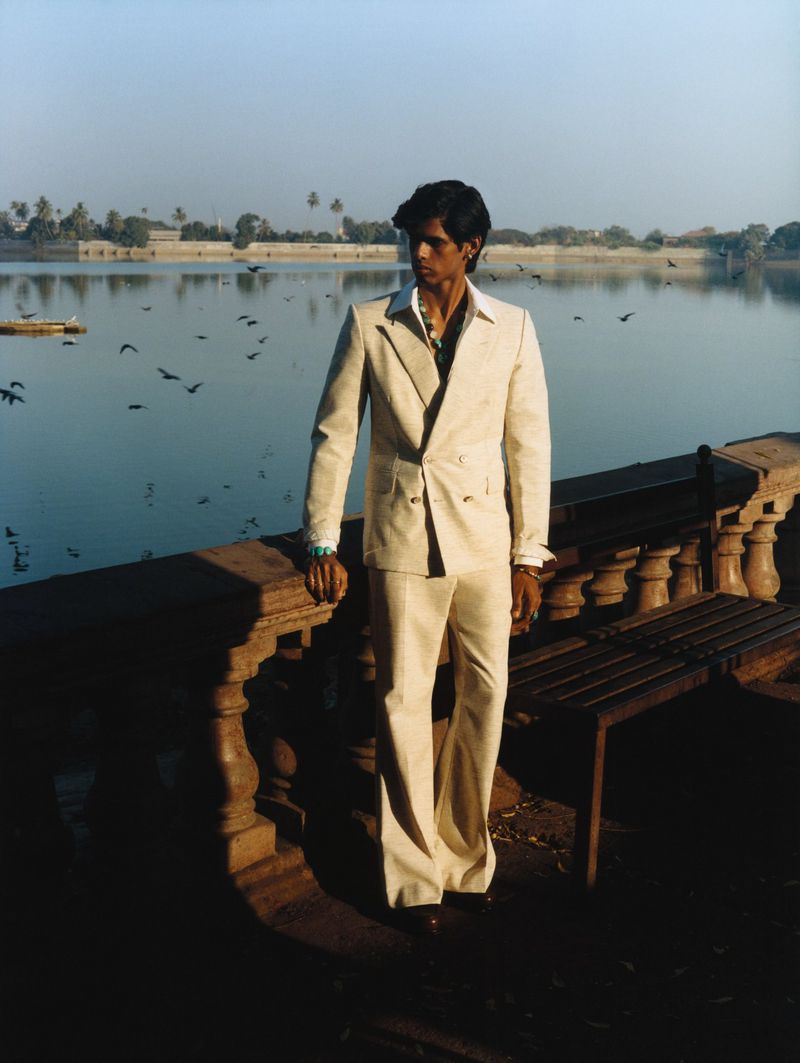 Man wearing a cream double-breasted suit standing by a stone balustrade overlooking a lake with birds in flight