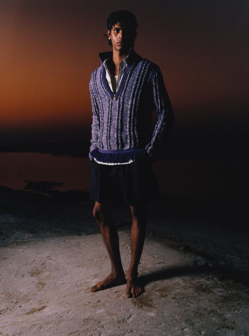 Man stands barefoot on shore in textured purple knit top and dark shorts, lit from below against a dark sky with faint orange glow.