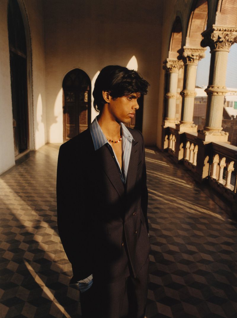 A man in a dark suit standing on a colonnaded balcony with sunlight streaming across the tiled floor.