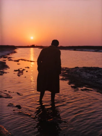 A silhouetted person wearing a long coat standing in shallow water during sunset.