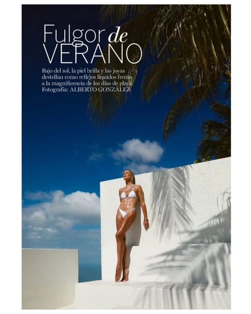 Woman in white bikini stands poolside on a sunny day. Palm tree fronds cast shadows on white wall. Blue sky with light clouds.