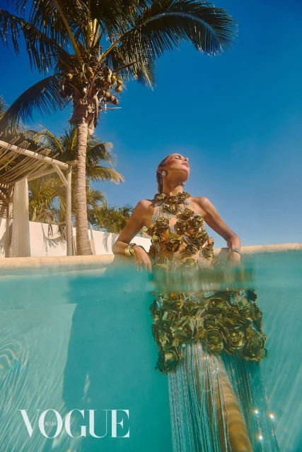 A woman in a floral dress stands in turquoise water, half-submerged, with palm trees and a clear blue sky in the background.  