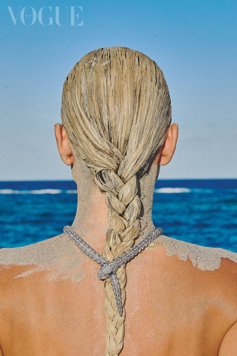 Blonde woman with braid and snake necklace on sandy beach looking at ocean under blue sky; Vogue logo in corner.