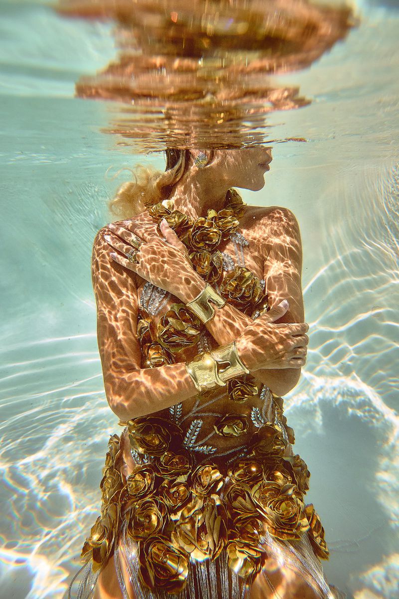 Underwater shot of a woman in an elaborate gold dress and jewelry with reflective water patterns on her skin in turquoise water.