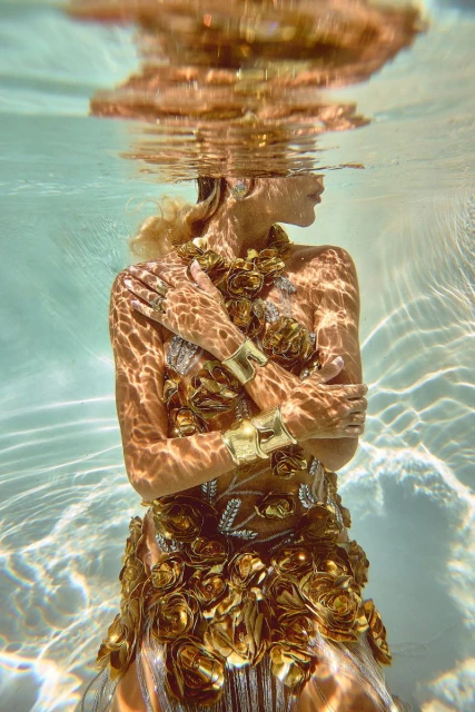 Underwater shot of a woman in an elaborate gold dress and jewelry with reflective water patterns on her skin in turquoise water.