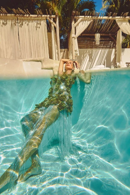 Adult woman in a green floral dress relaxing in a swimming pool, featuring clear blue water and a tropical cabana background.