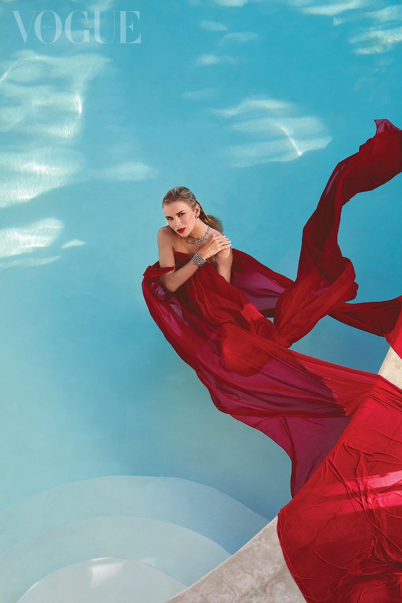 Blonde woman in a red gown poses in water near a concrete structure with Vogue magazine logo visible in the upper left corner.