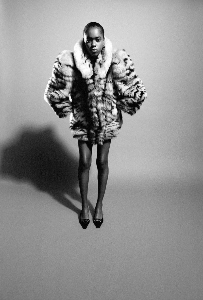 Model in patterned fur coat and black shoes standing against a grey studio backdrop