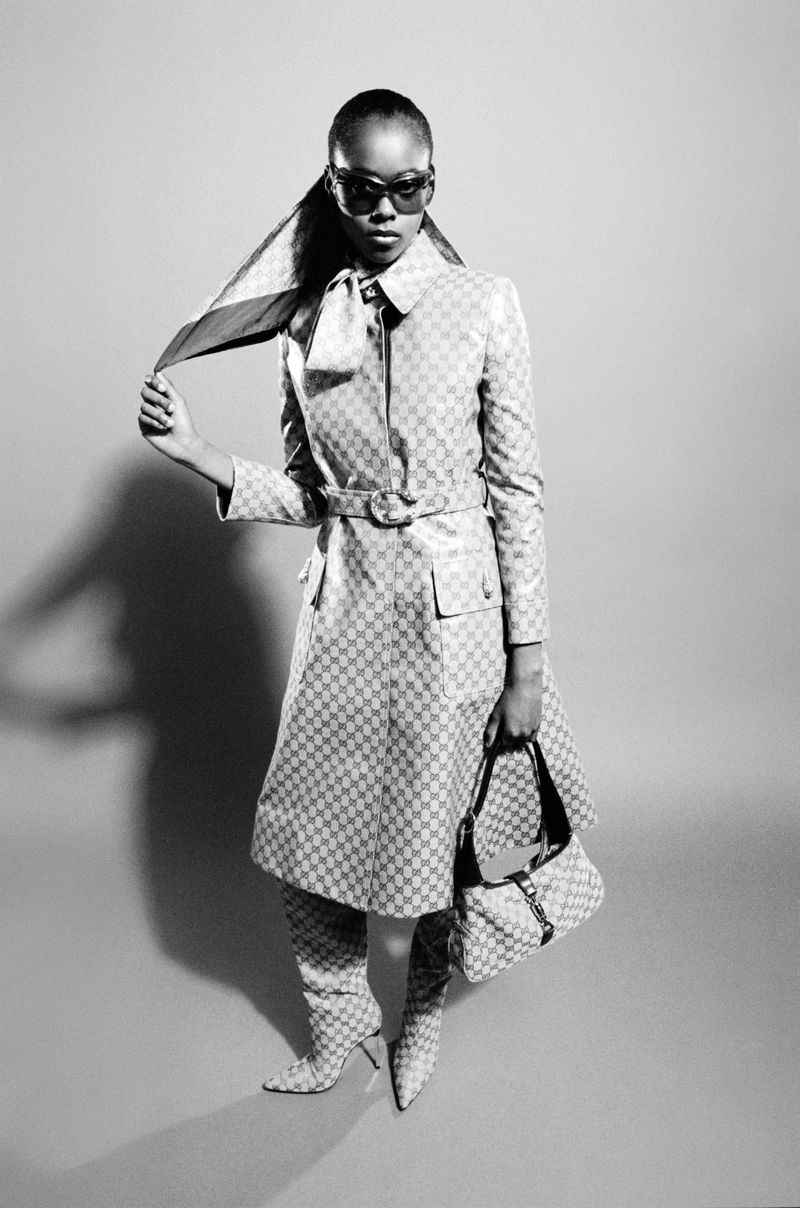 Model wearing a patterned trench coat, knee-high boots, and sunglasses holding a patterned handbag against a grey backdrop
