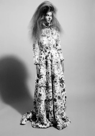 Model with voluminous hair wearing a long patterned dress standing against a plain background