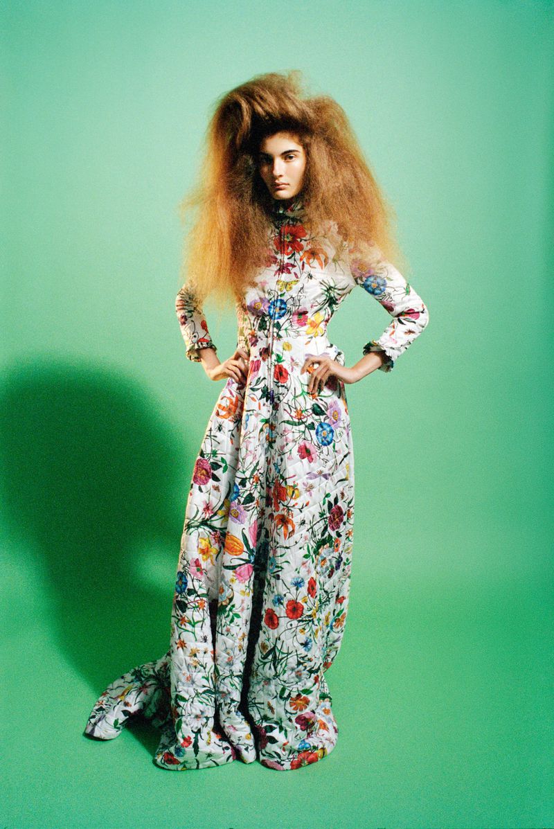 Model with voluminous hair wearing a long floral print dress standing against a green backdrop