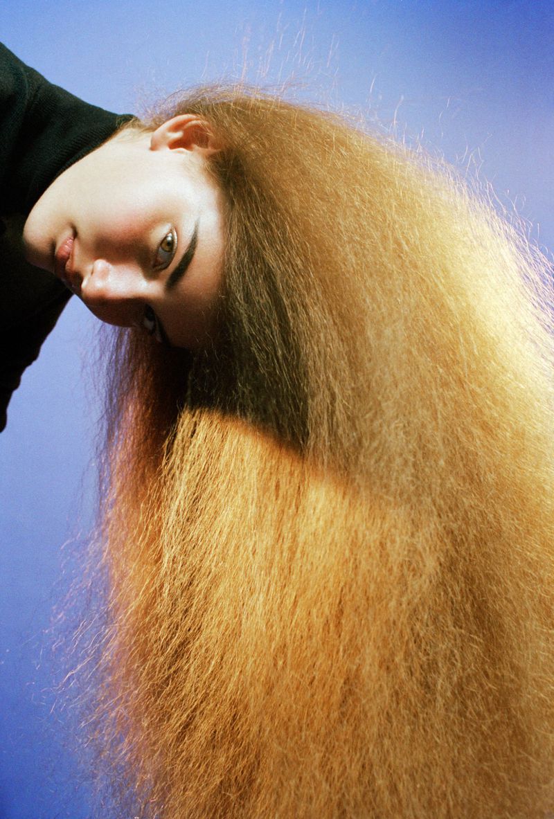 Portrait of a model with voluminous long blonde hair cascading downward against a plain blue background.