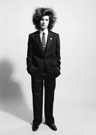 Model with voluminous hair wearing a dark suit and tie standing against a plain light background