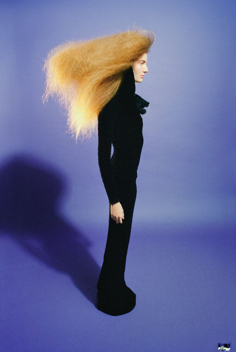 Model in profile wearing a black turtleneck and long skirt with voluminous blonde hair blowing horizontally against a blue backdrop