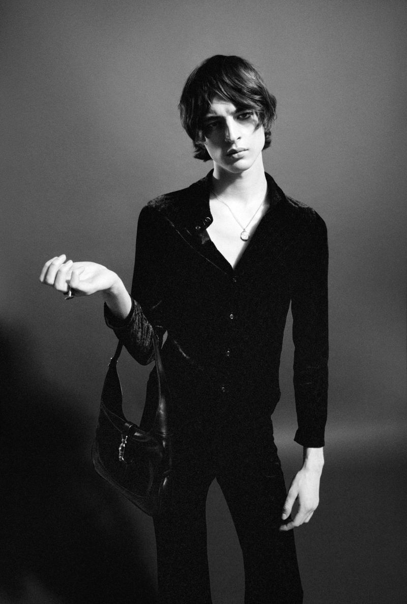 Young man in black shirt holding a black leather handbag against a grey seamless studio background.
