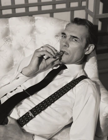 Monochrome portrait of a man in a white shirt and patterned suspenders seated on a tufted chair.