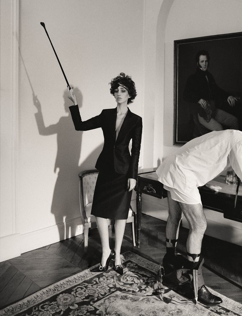 Monochrome editorial image featuring a woman in a dark skirt suit holding a whip standing next to a man in white underwear