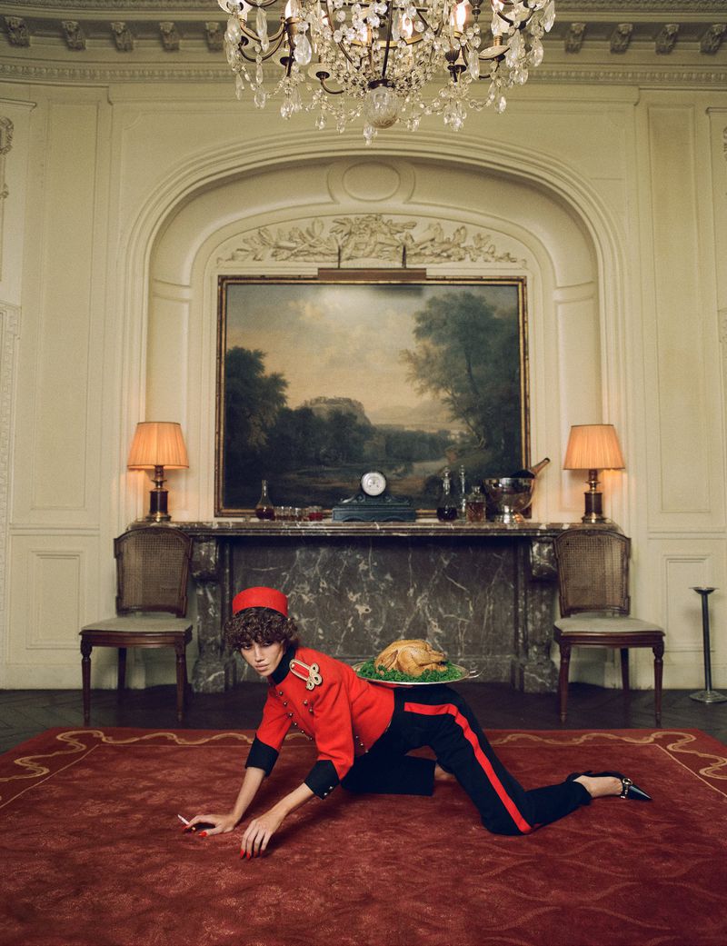 Model in red bellhop uniform kneeling on a red rug holding a platter with a roasted turkey in an ornate room.