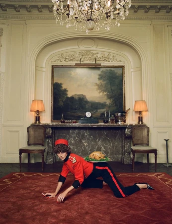 Model in red bellhop uniform kneeling on a red rug holding a platter with a roasted turkey in an ornate room.