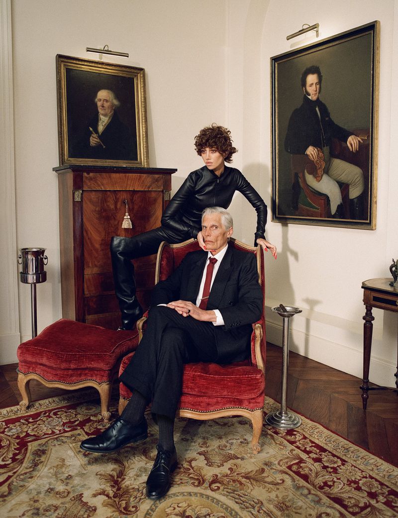 An elderly man seated in a red armchair with a woman in a black leather jumpsuit posed behind him in a room with framed portraits